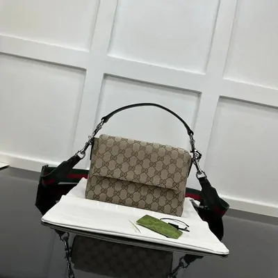 A small flap shoulder bag in tan monogram-printed canvas with a dark leather top handle, propped on a white stand against a panelled grey wall