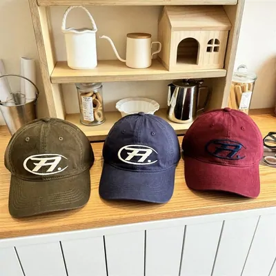Three corduroy baseball caps lined up on a kitchen counter, olive green, navy and dark red, each with the same embroidered motif on the front panel
