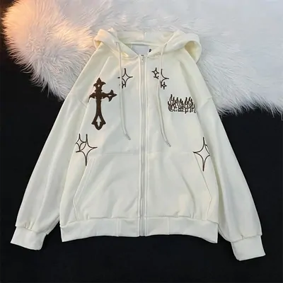 A cream zip-through hoodie laid flat on a dark surface, embroidered cross, four-point stars and a flame motif across the chest, drawstring hood