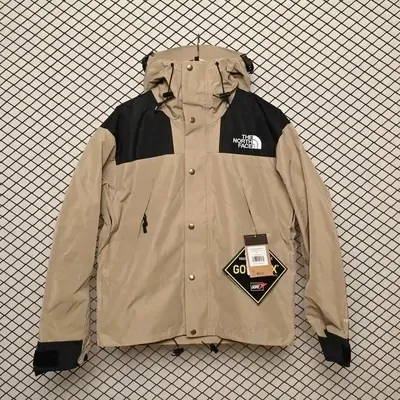 A hooded shell jacket laid flat on dark mesh, sand-coloured body with black shoulder and hem panels, zip pockets and a paper hang tag still attached