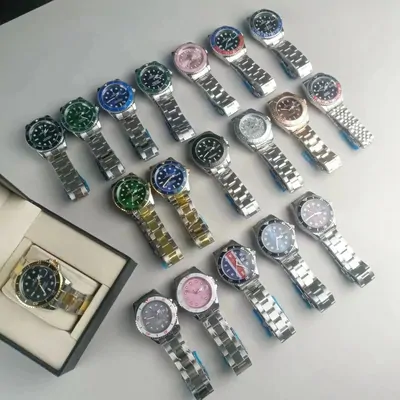 About twenty steel-bracelet dive-style wristwatches laid out in rows on a grey surface, bezels in black, green, blue, pink and two-tone gold, with one more watch sitting in an open presentation box at the left